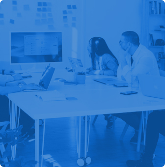 A group of people sit around a table in an office setting, engaging with laptops and a large monitor. The room has shelves and sticky notes on the wall. The image has a blue tint overlay.